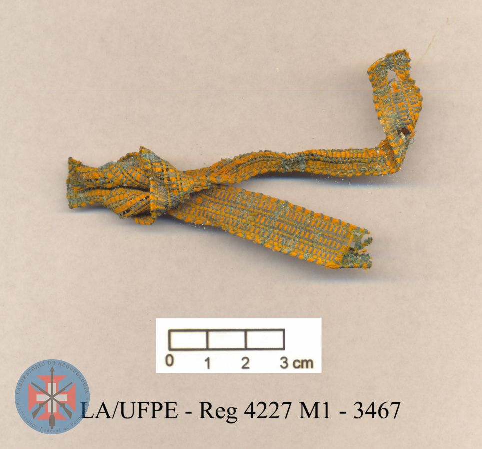 Fragmento de fita de medalha, em tecido verde e laranja. Material associado a sepultamento.